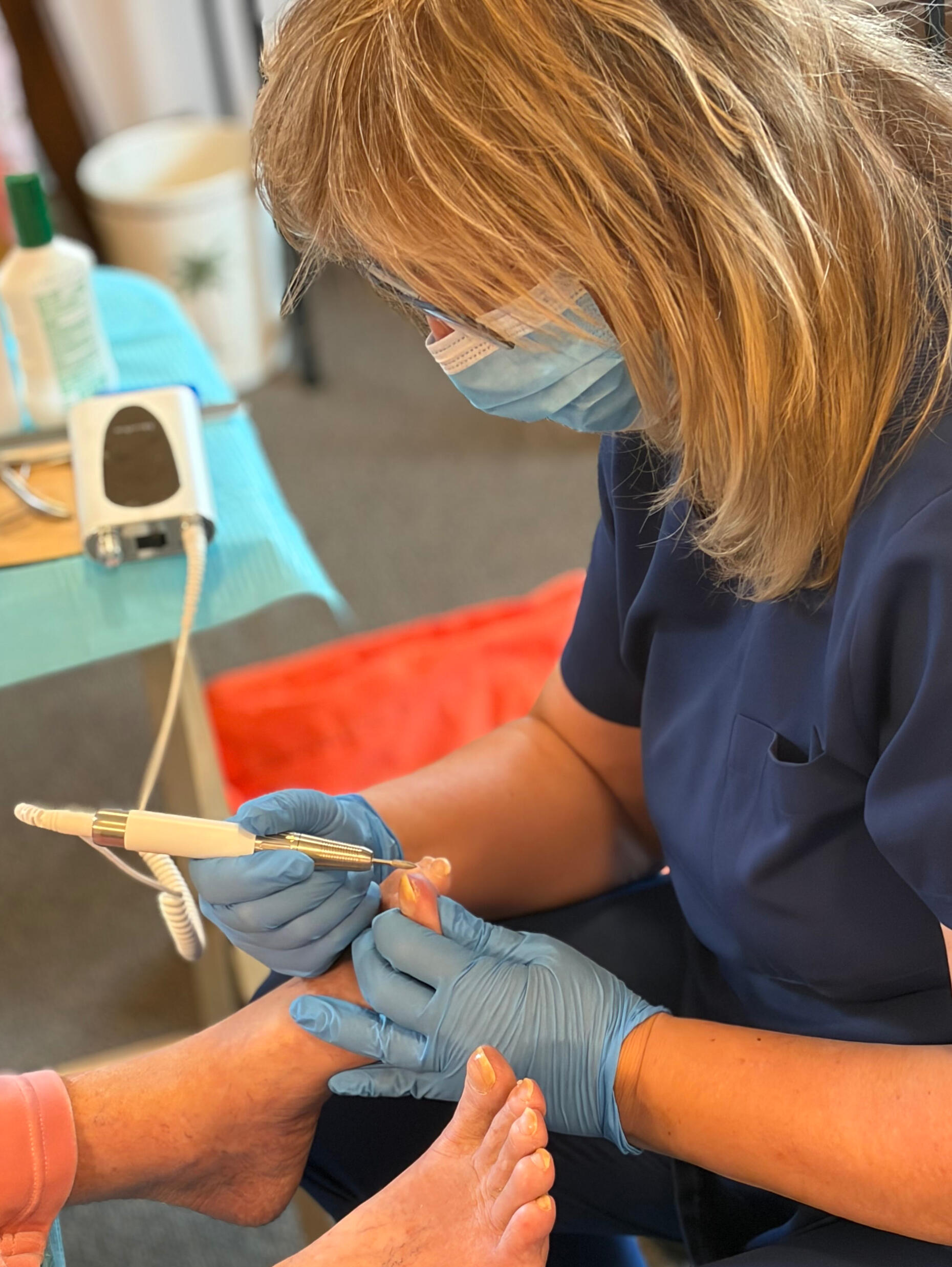 Registered nurse providing in-home toenail care using a professional nail sanding tool.